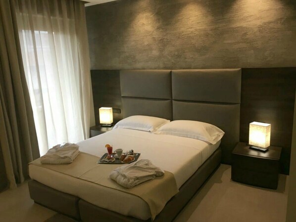 A modern hotel room with a large bed, two bedside tables with lamps, a tray with breakfast items, and a view of the outdoors through sheer curtains. - Plus Welcome Milano (Milan)