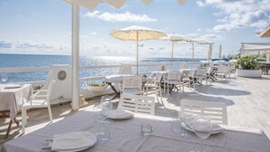 Un área de comedor al aire libre con mesas y sillas, sombrillas blancas y vista al mar.