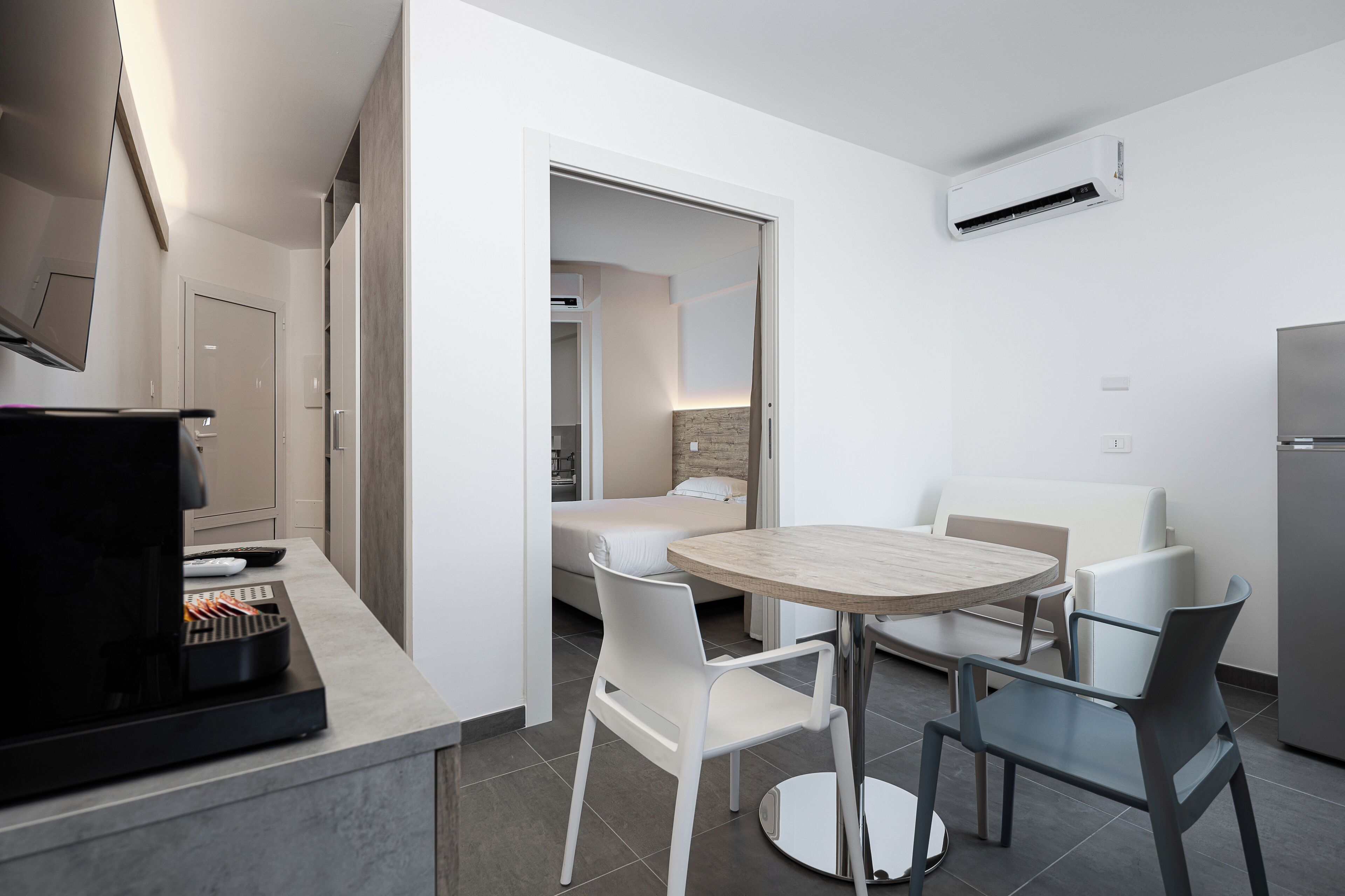 A modern kitchen with a coffee machine, a dining table with chairs, and a view into a bedroom.
