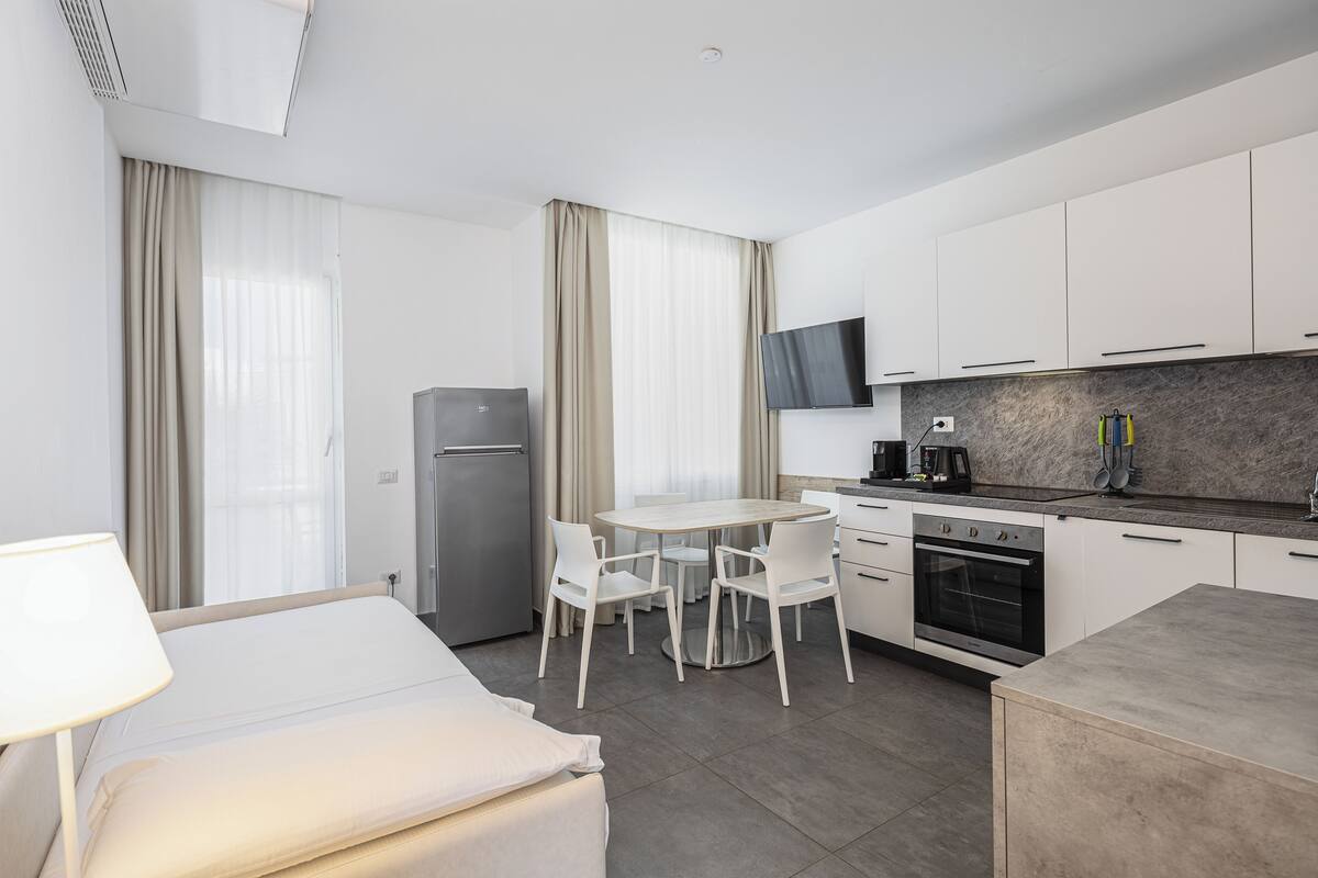 Una habitación de hotel moderna con cama, escritorio, una mesa de comedor con sillas, y una cocina que incluye estufa y televisor sobre la encimera.