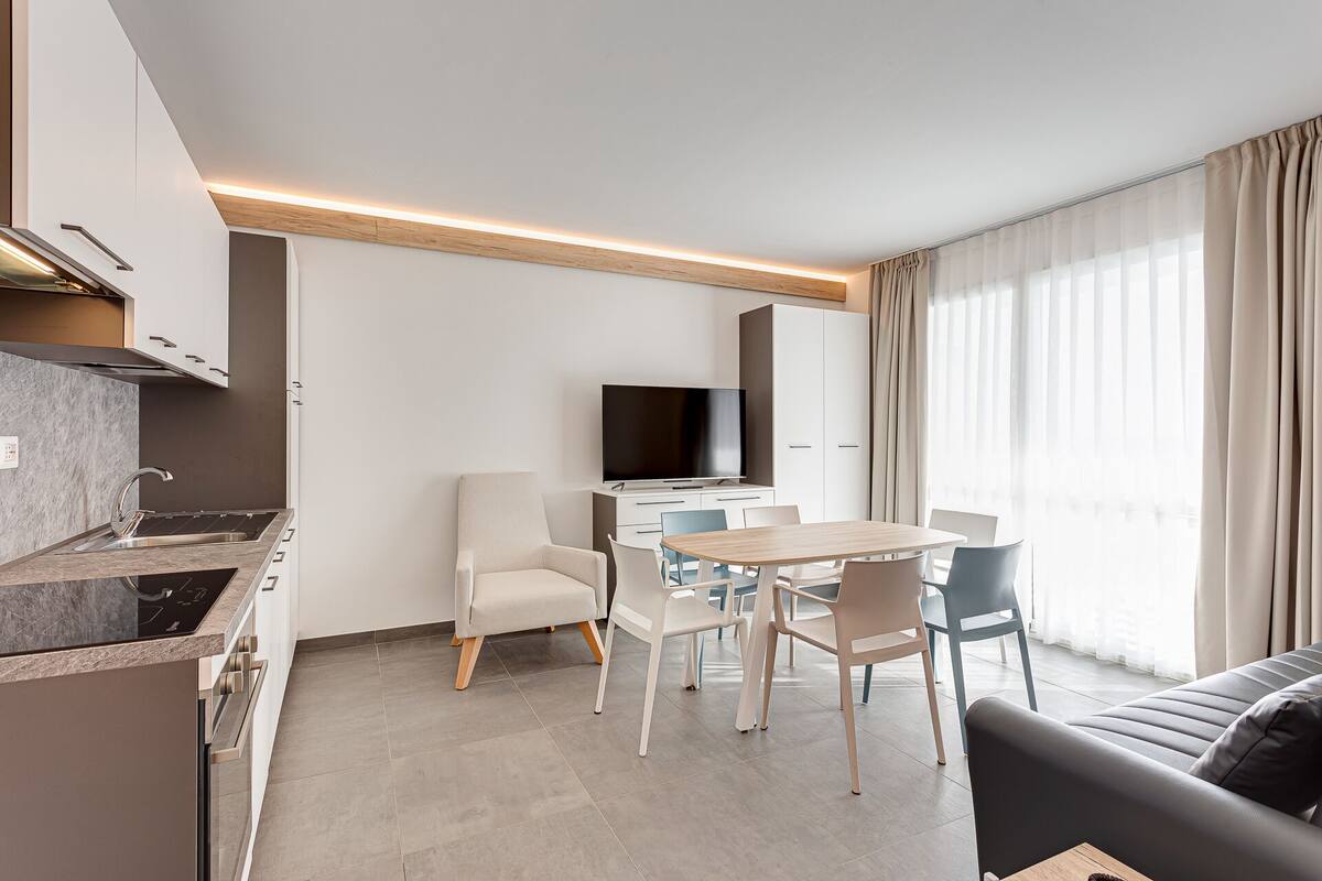 Habitación de hotel moderna y bien iluminada, con área de cocina, mesa de comedor con sillas, televisor de pantalla plana y sofá.
