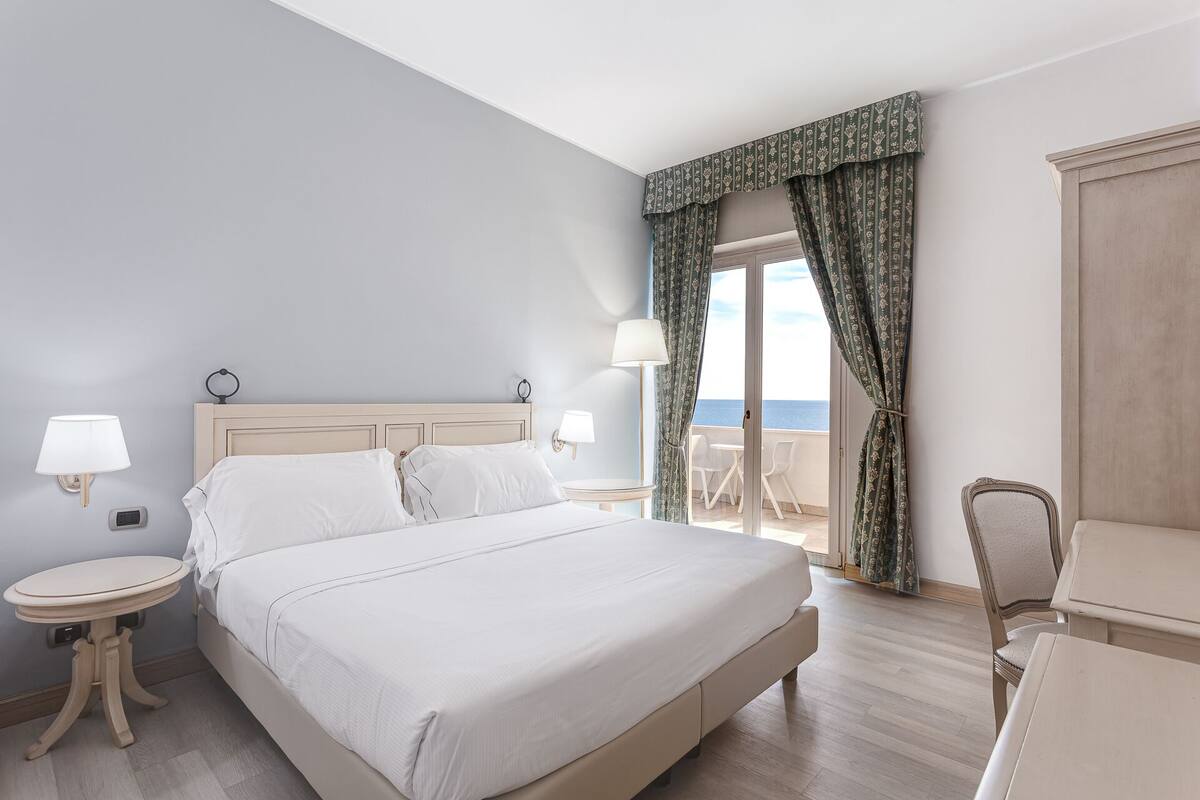 Habitación de hotel con una cama grande, dos mesitas de noche, una silla y vistas al mar.