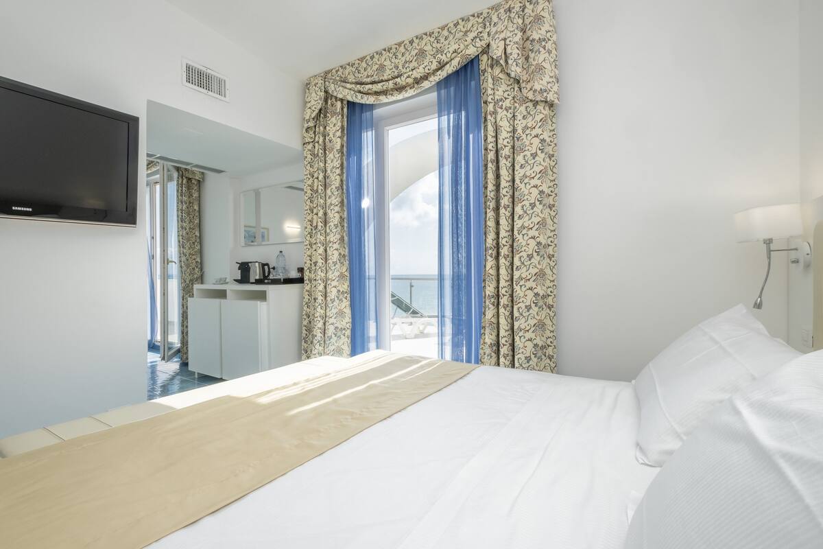 Un cuarto de hotel con cama, televisor montado en la pared, ventana con cortinas y vista al mar.