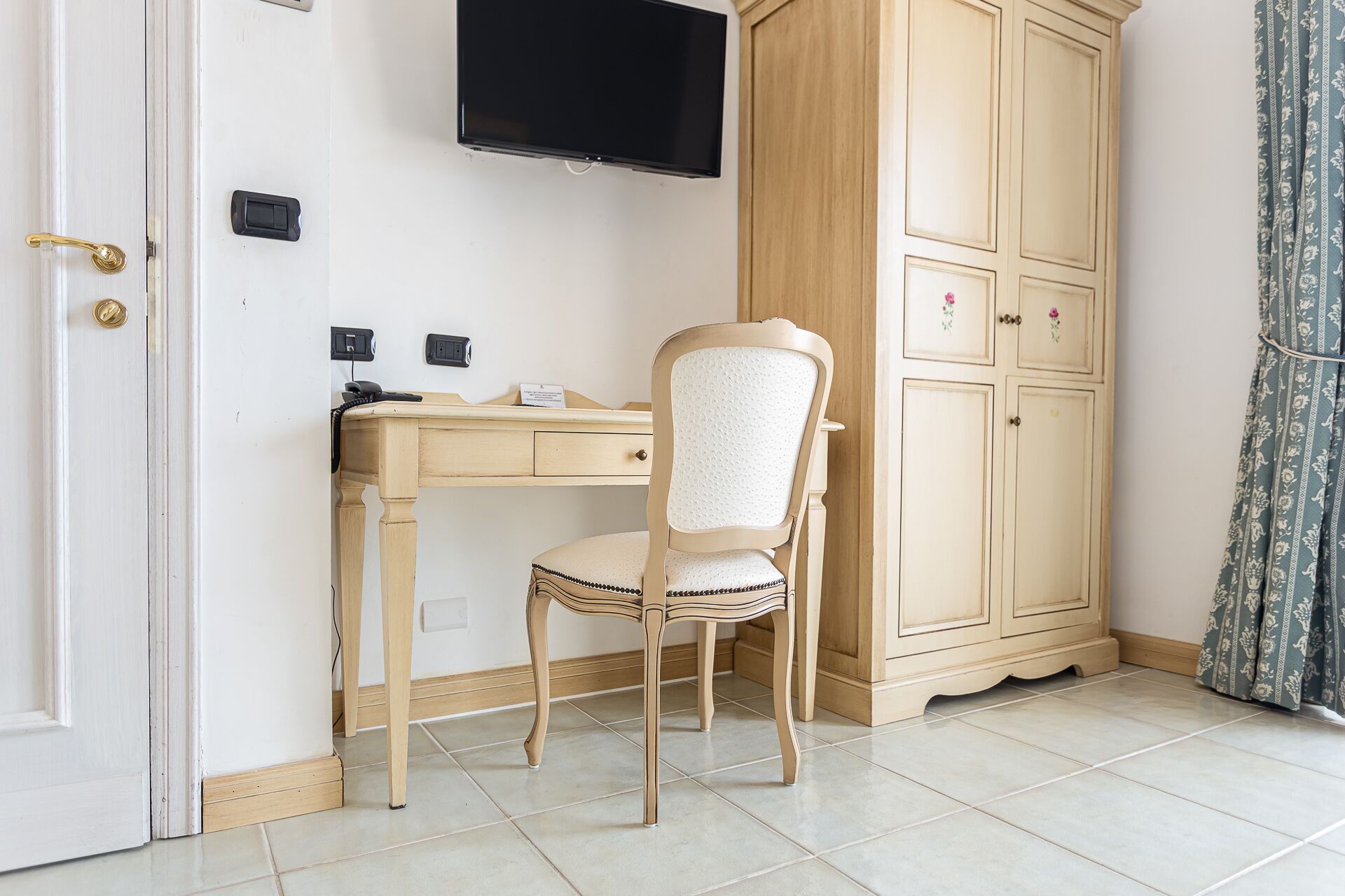 Um quarto com uma escrivaninha de madeira, uma cadeira, uma televisão na parede, um guarda-roupa e uma porta com puxador de latão.