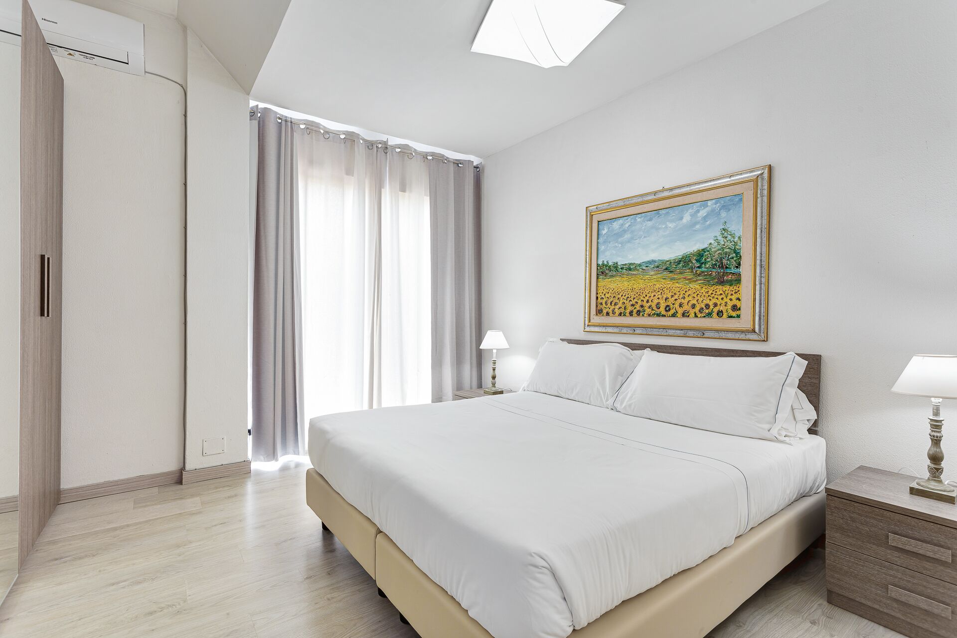 A hotel room with a bed, a nightstand, a lamp, a painting, and a window with curtains.