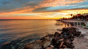 Una puesta de sol costera con un muelle que se adentra en el agua, costa rocosa y edificios al fondo.