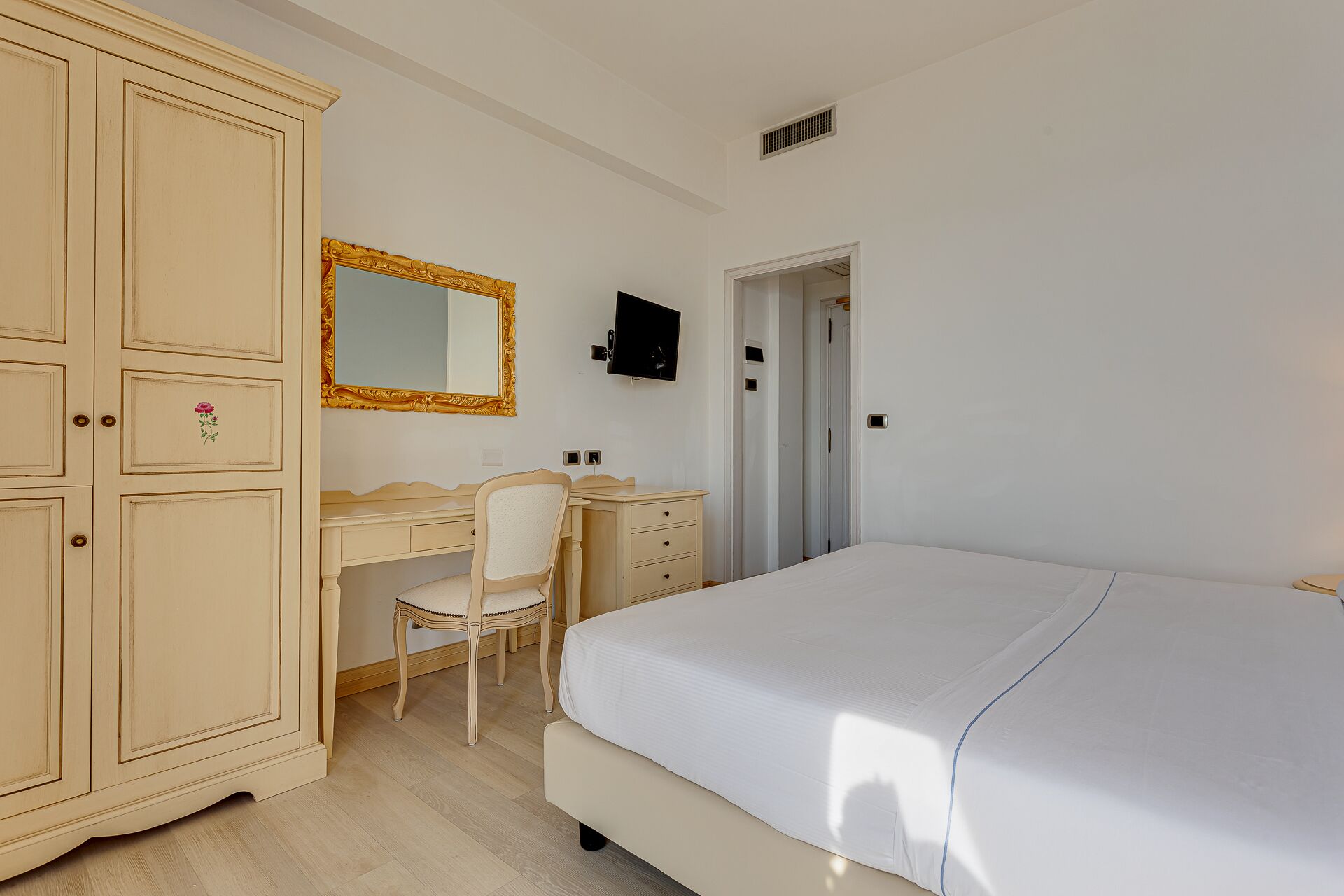 A bedroom with a bed, a desk with a chair, a wardrobe, a television, and a door leading to another room.