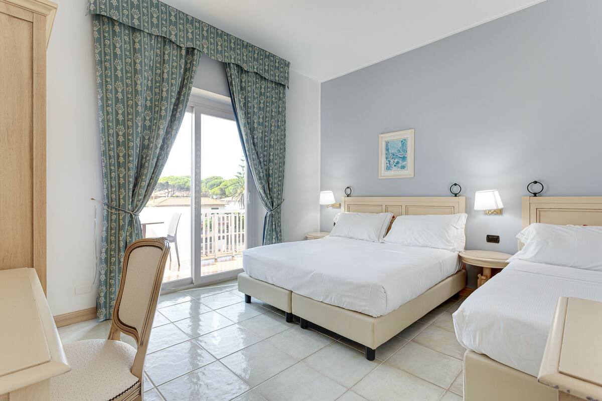 Habitación de hotel con dos camas, una silla, un balcón con cortinas y un cuadro en la pared.