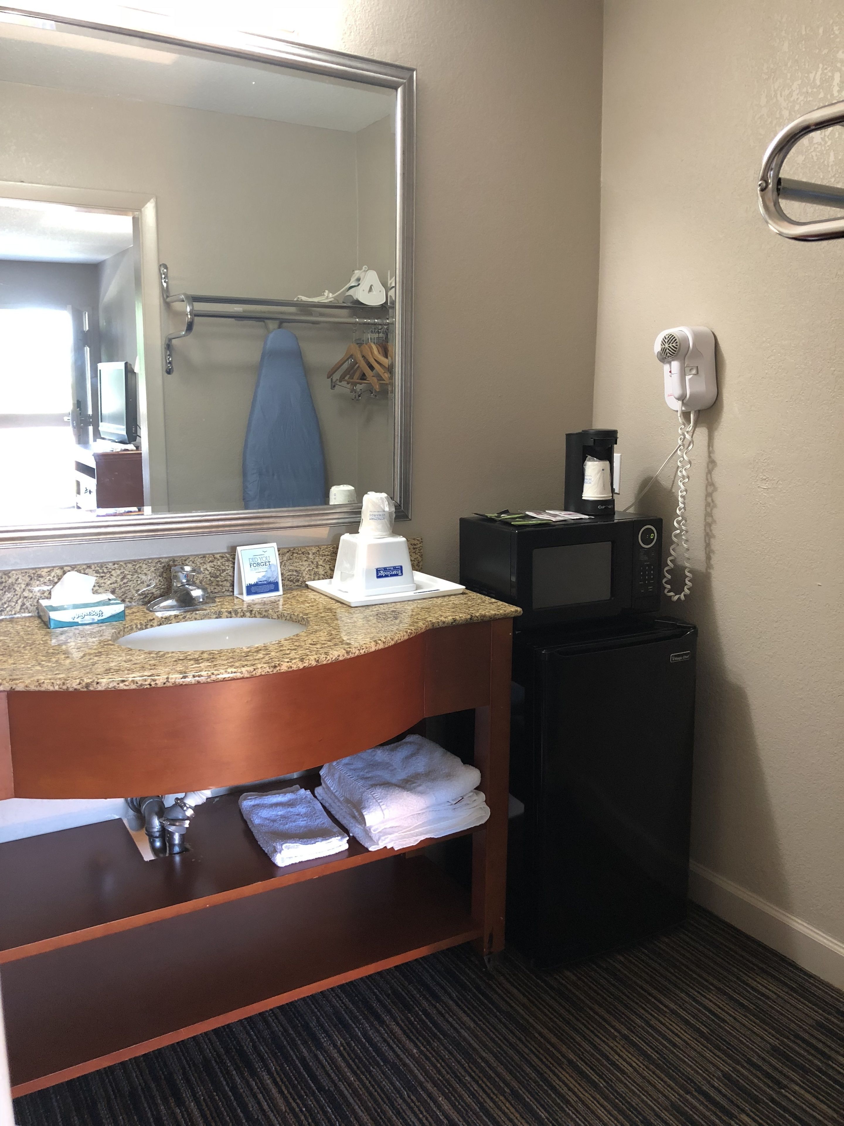A hotel bathroom with a granite countertop sink, a mirror, a hairdryer, and a small refrigerator.
