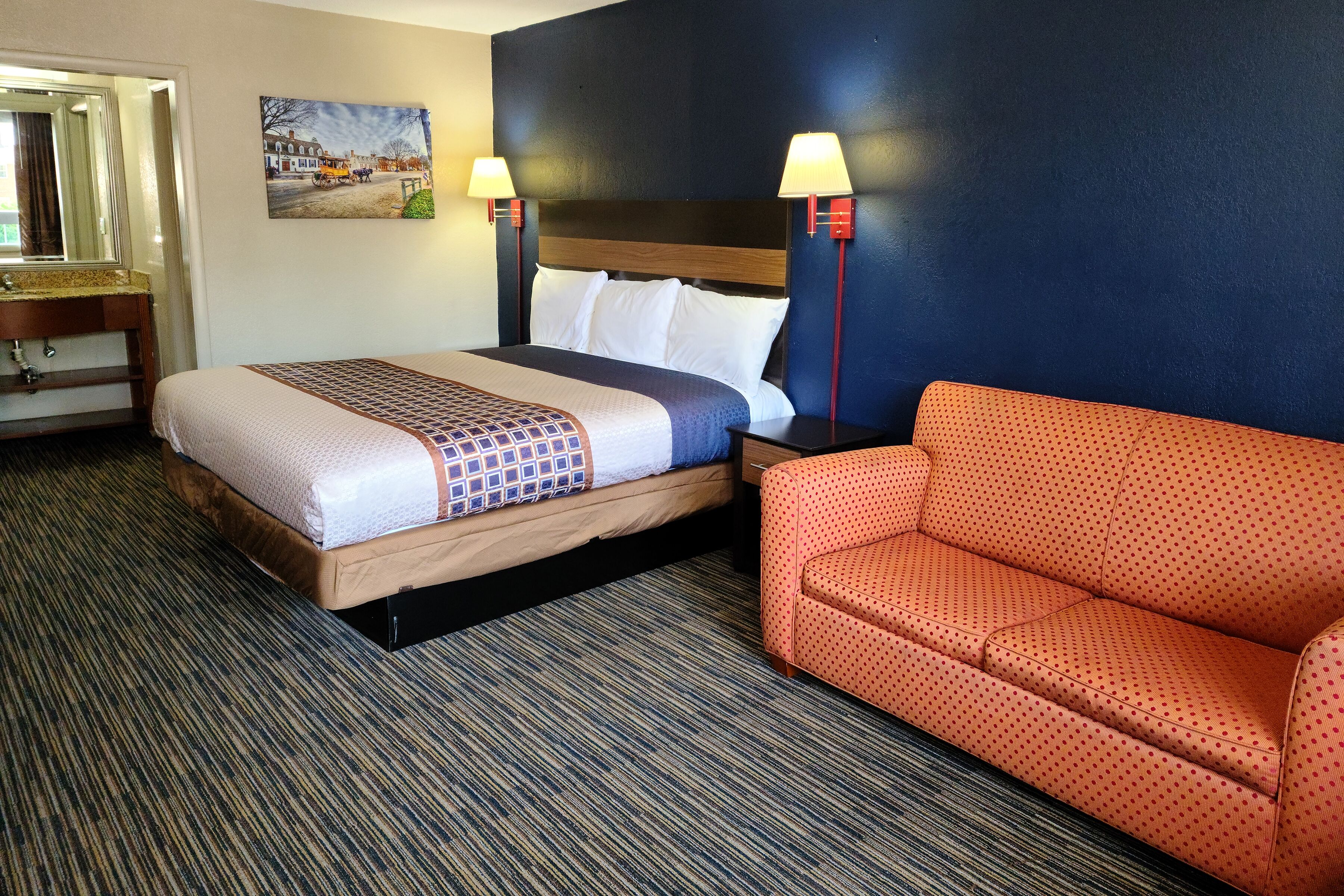 A hotel room with a bed, a sofa, a painting, and a nightstand with lamps.