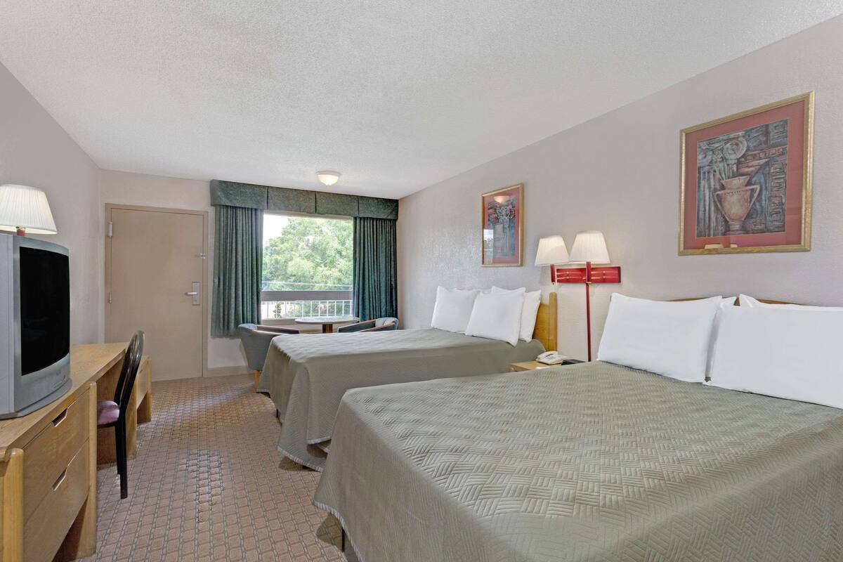A hotel room with two beds, a desk with a TV, a window with curtains, and a painting on the wall.