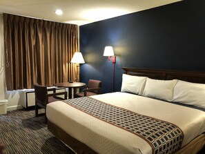 A hotel room with a large bed, a desk, two chairs, a lamp, and a window with curtains. - Travelodge by Wyndham Williamsburg Colonial Area (Williamsburg)