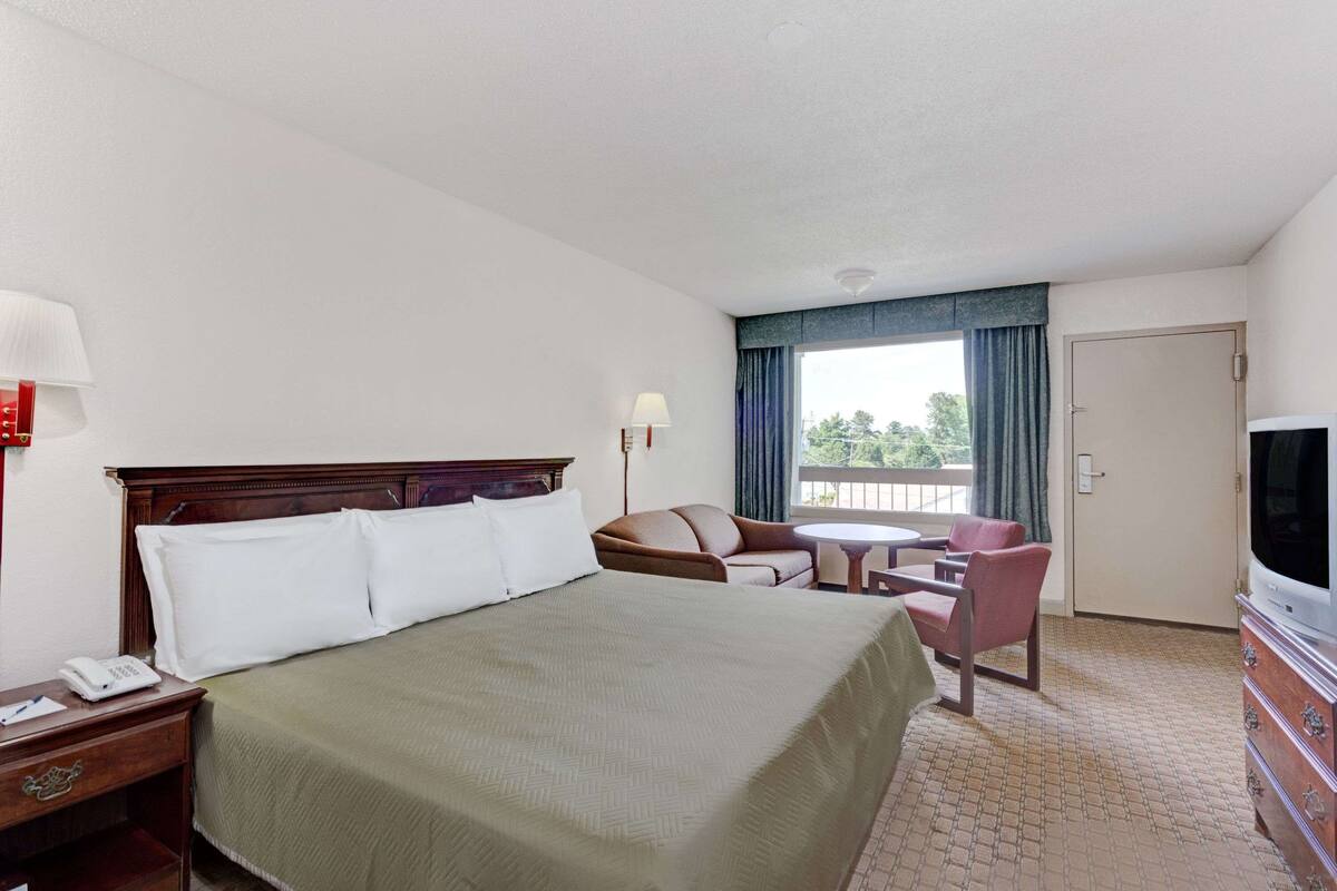 A hotel room with a large bed, a desk with a phone, a TV, a small table with chairs, and a balcony with curtains.