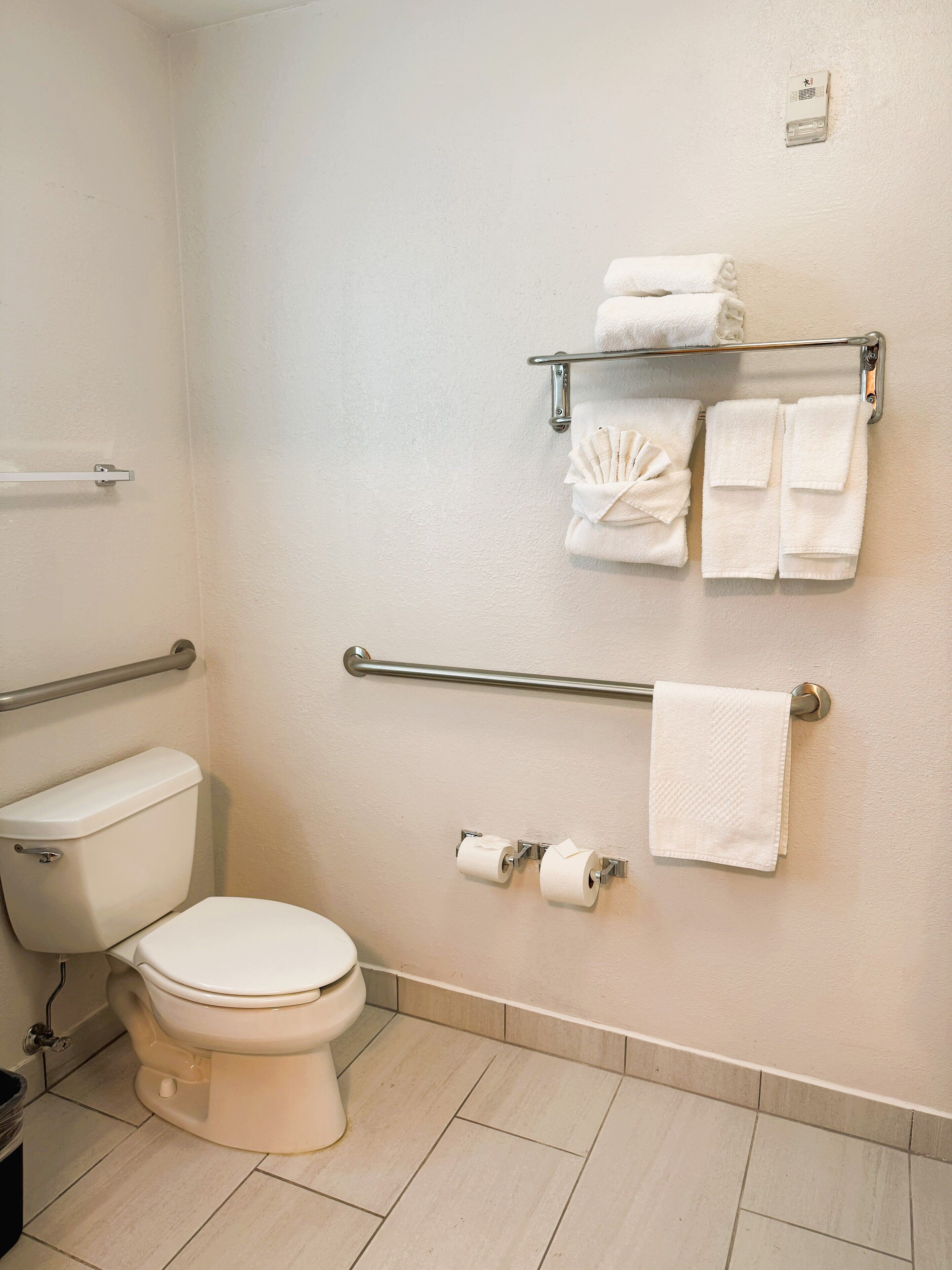 A bathroom with a toilet, grab bars, a towel rack with towels, and toilet paper.
