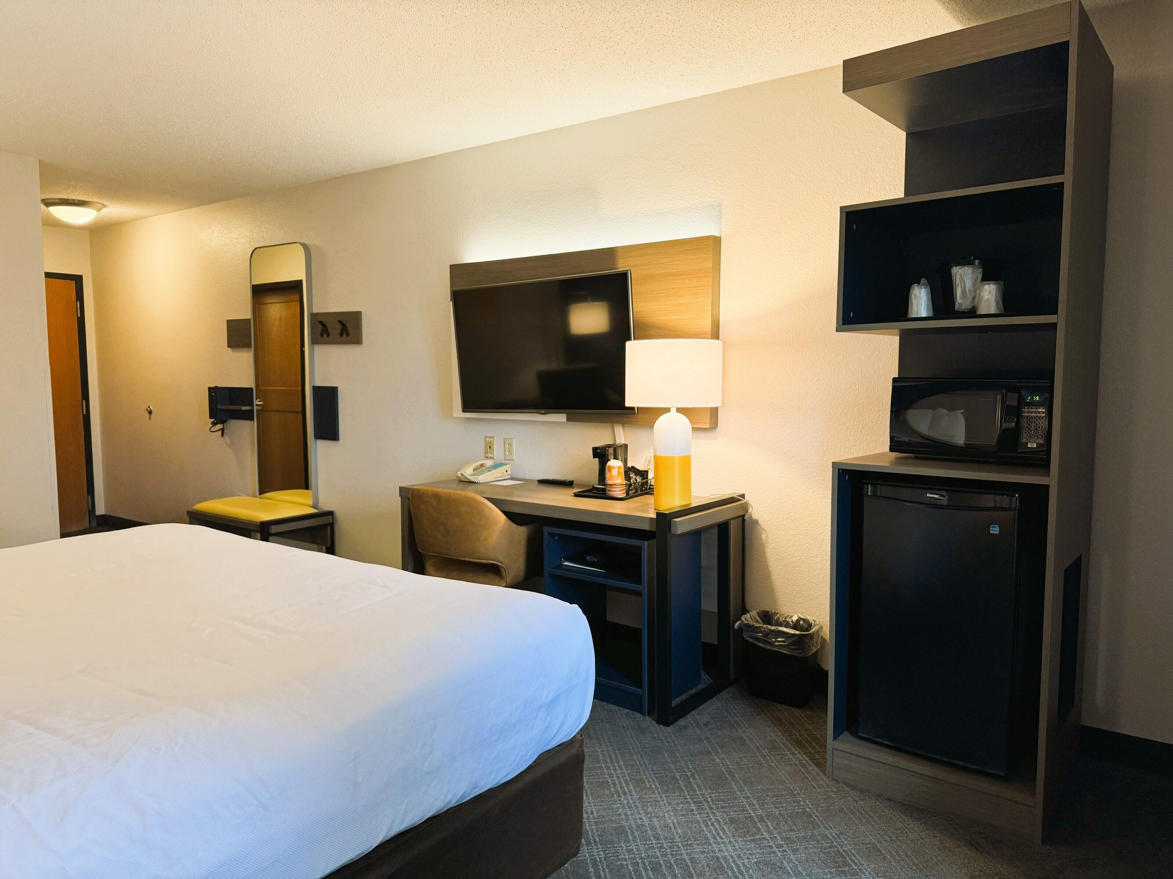Habitación de hotel con cama, escritorio, silla, televisor, lámpara, microondas y refrigerador pequeño.