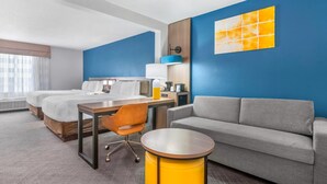 A hotel room with two beds, a desk, a chair, a sofa, and a round yellow table.