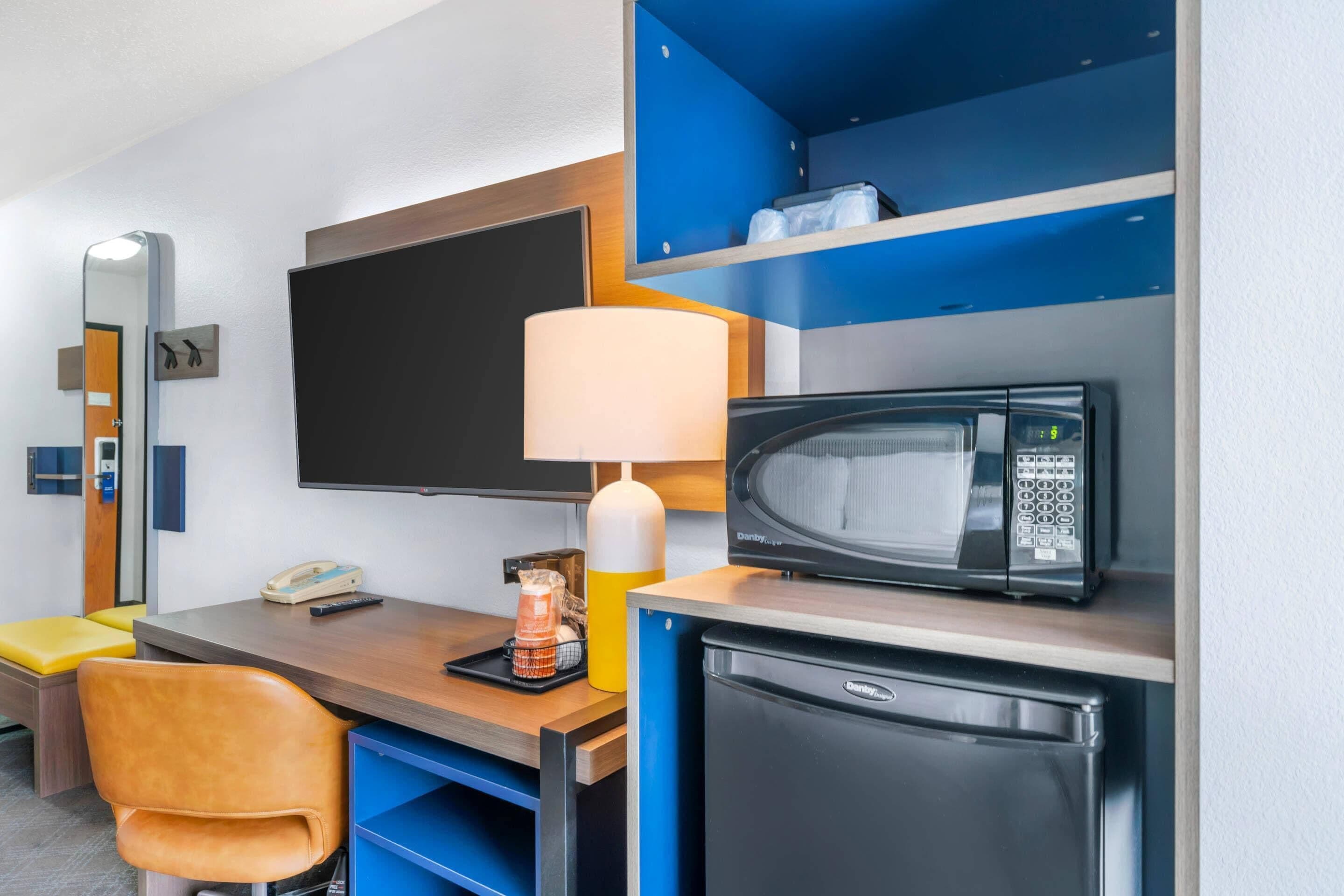 Habitación de hotel con escritorio, microondas, refrigerador, lámpara, teléfono, silla y televisor.