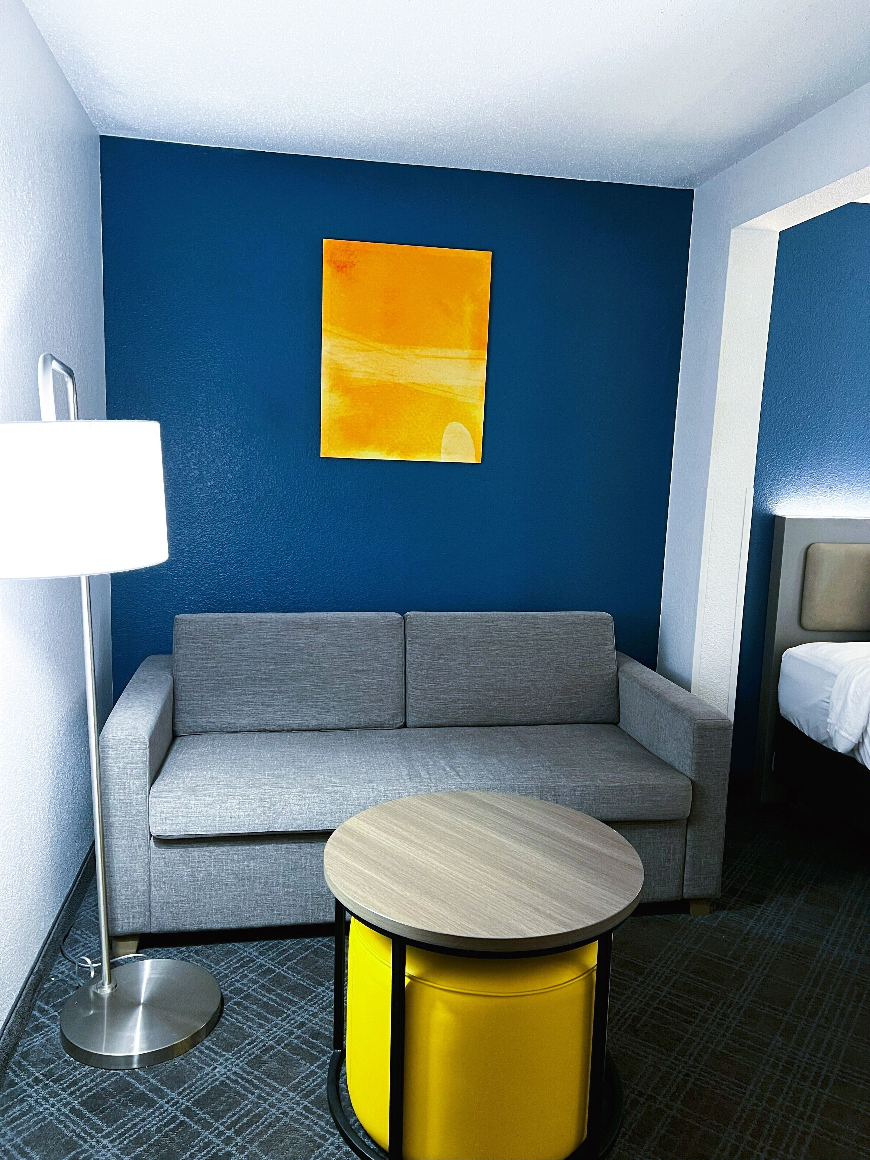 Ein Hotelzimmer mit grauem Sofa, einem runden Holztisch, einem gelben Hocker, einer weißen Lampe und einem Bild an der Wand.