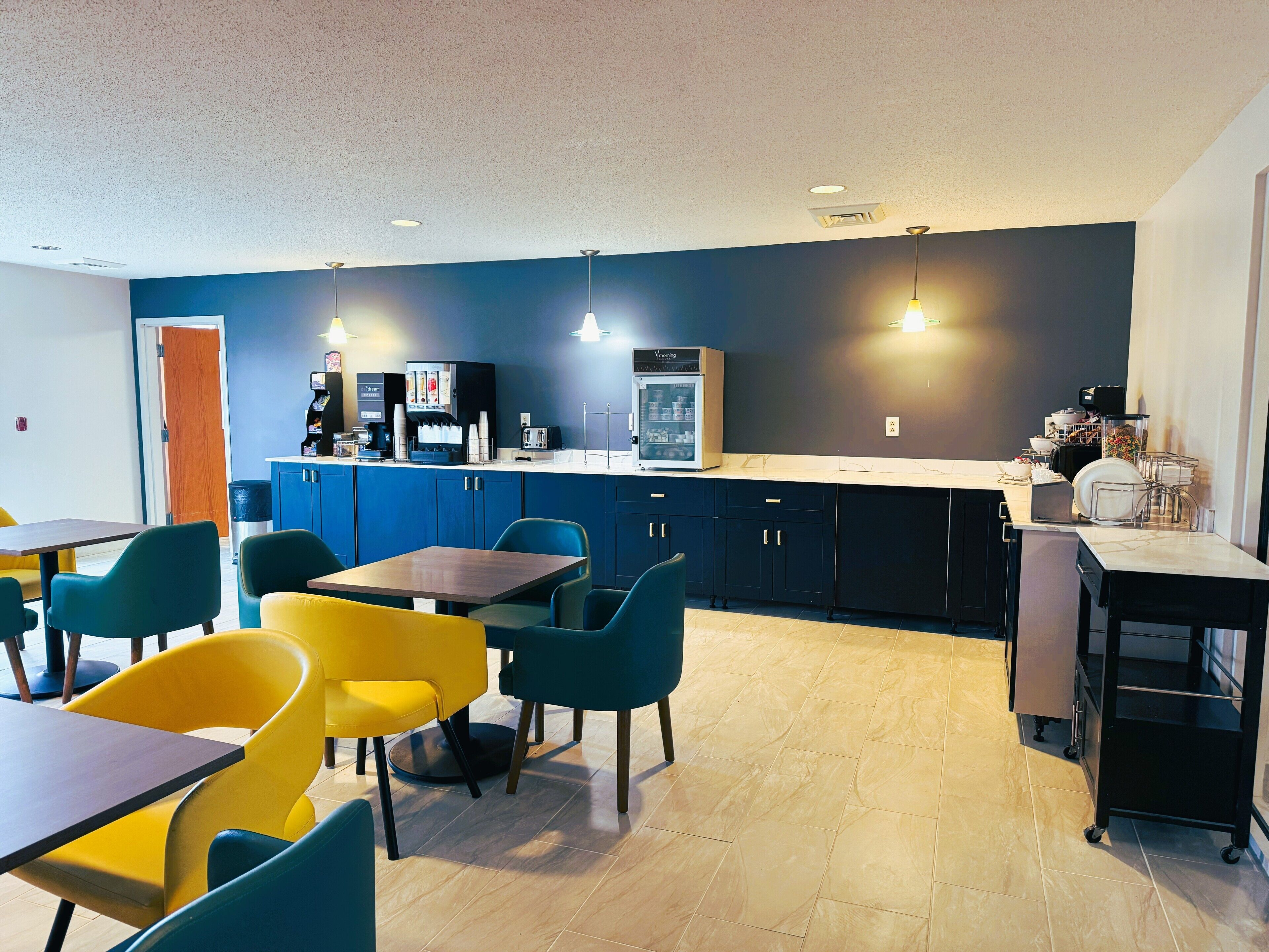 Un espace cuisine moderne équipé d’une machine à café, d’un réfrigérateur et d’un comptoir où sont disposés divers objets. On aperçoit des chaises et une table au premier plan.