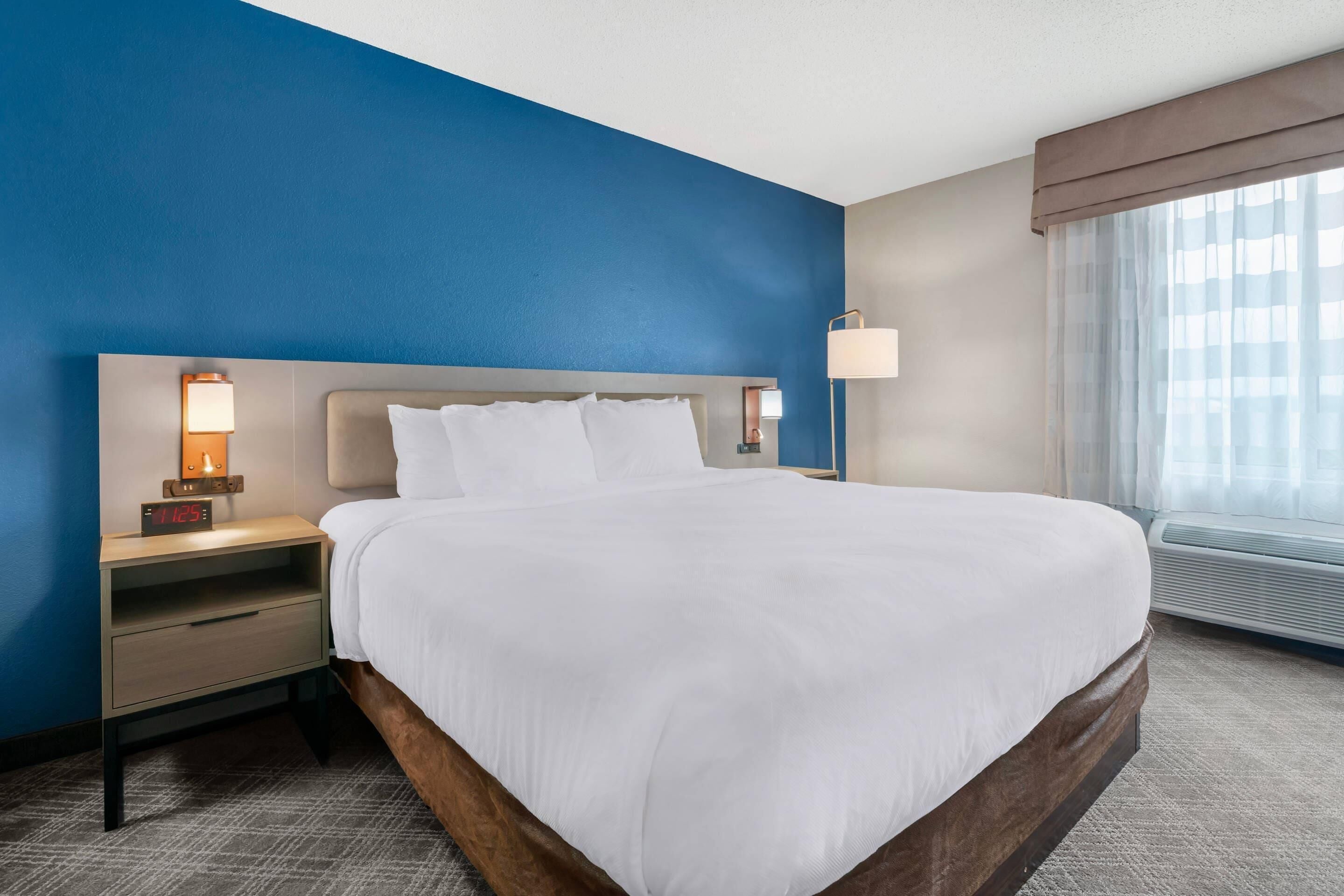 A hotel room with a large bed, a nightstand with a lamp, a wall clock, and a window with curtains.