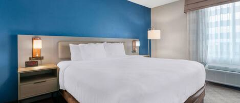 A hotel room with a large bed, a nightstand with a lamp, a wall clock, and a window with curtains.