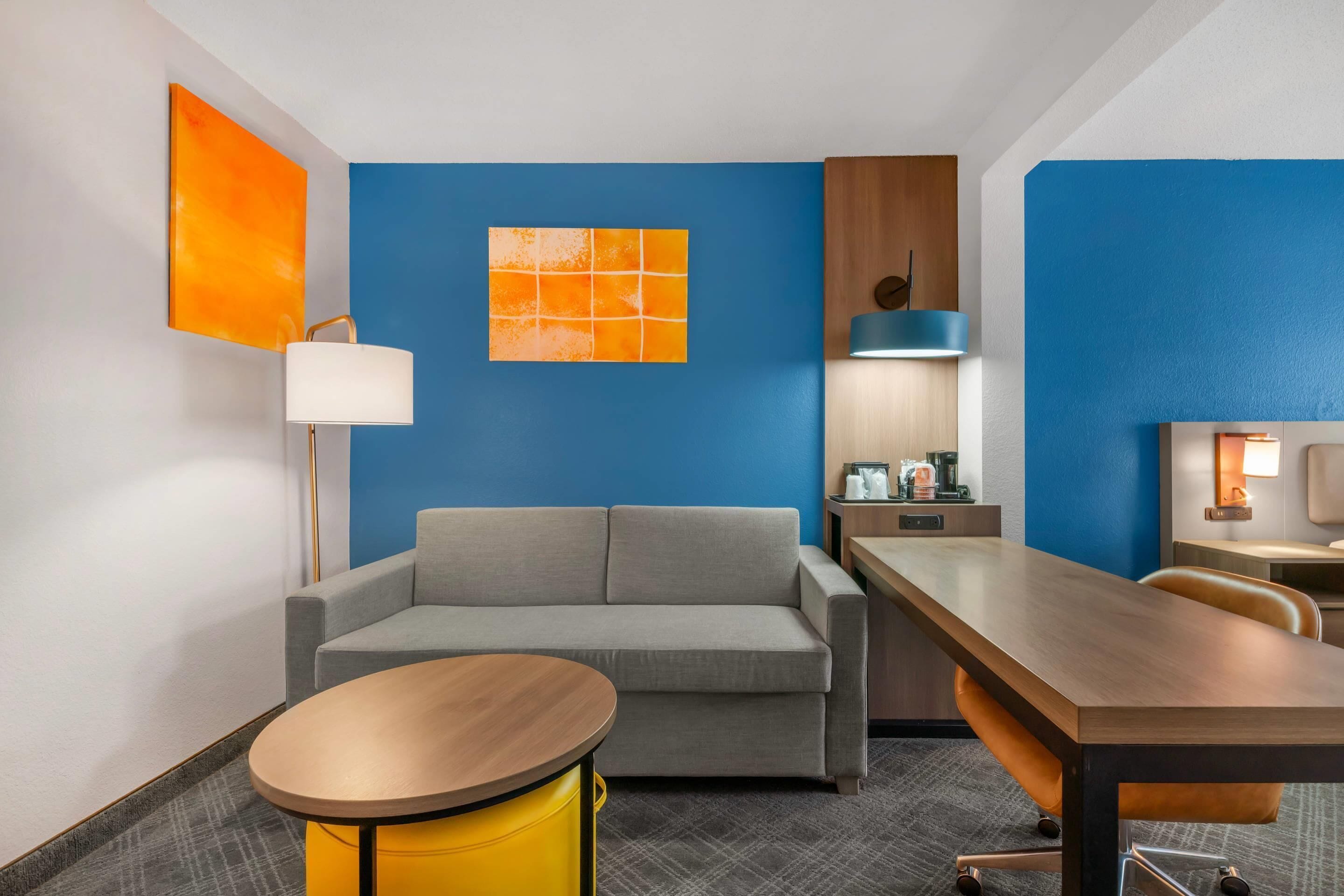 A modern hotel room with a gray sofa, a wooden dining table, a yellow side table, and a blue accent wall.