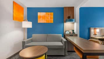 A modern hotel room with a gray sofa, a wooden dining table, a yellow side table, and a blue accent wall.