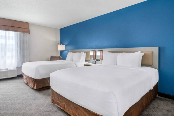 A hotel room with two beds, a large window with curtains, a bedside table with a lamp, and a blue accent wall.