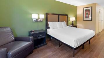 Habitación de hotel con cama, mesita de noche, lámpara, sillón y un cuadro en la pared.