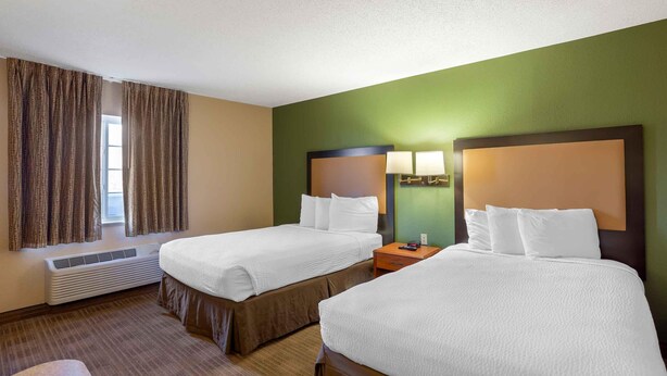 A hotel room with two beds, a window with curtains, a heater, and a nightstand with a lamp.