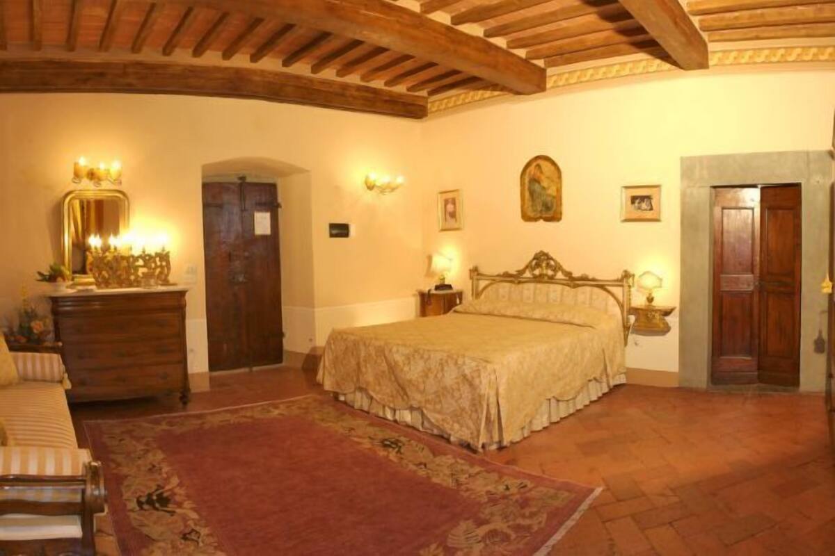 Un dormitorio con techo de madera, cama con colcha floral, cómoda, espejo y apliques de pared.