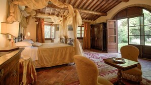 A bedroom with a large bed, wooden ceiling beams, a window with a view of greenery, and a small table with a bowl. - Relais Villa Baldelli (Cortona)