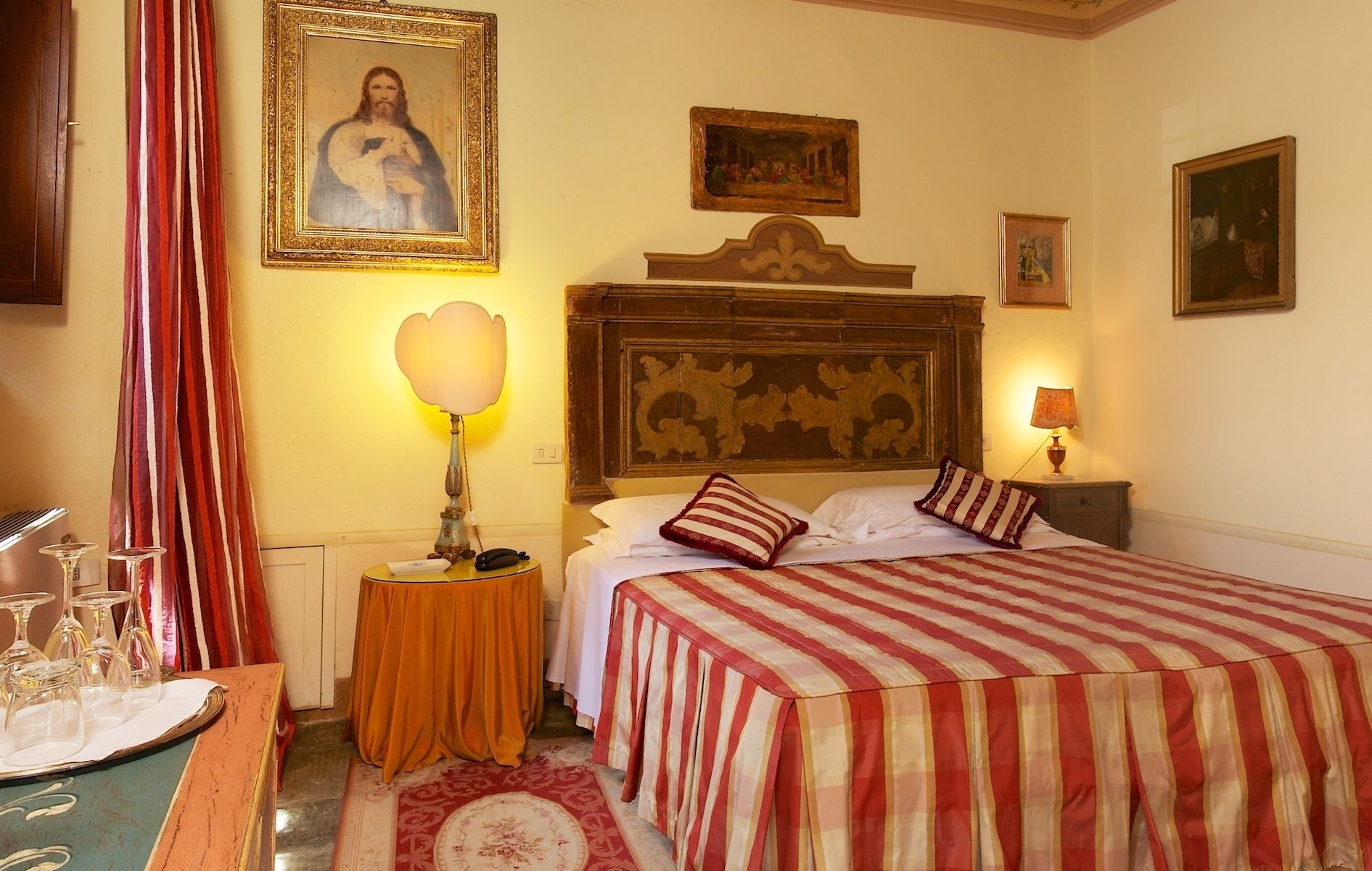 A hotel room with a bed, a nightstand, a lamp, a framed picture on the wall, and a table with glasses.