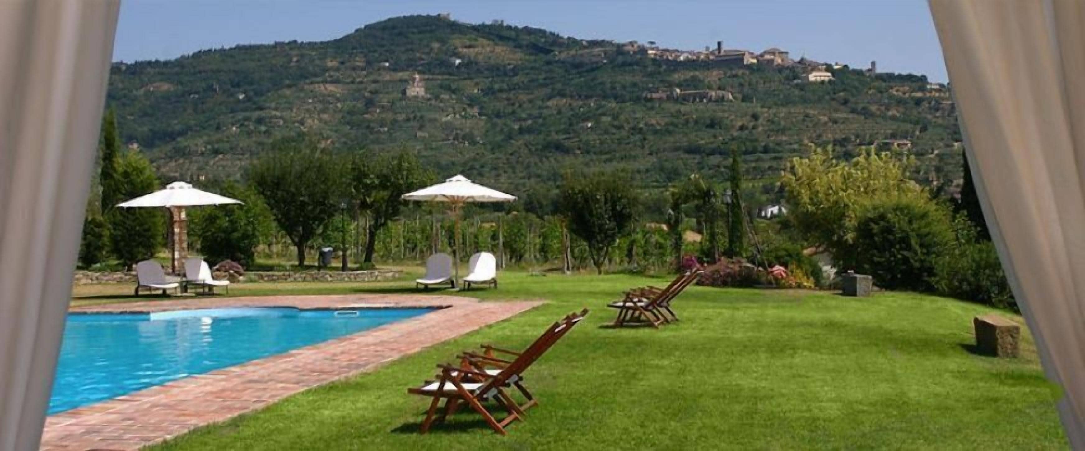 A pool area with lounge chairs and umbrellas, surrounded by a grassy lawn and a view of a hill with buildings.
