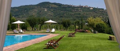 Un espace piscine doté de chaises longues et de parasols, entouré d’une pelouse verdoyante et offrant une vue sur une colline parsemée de bâtiments.