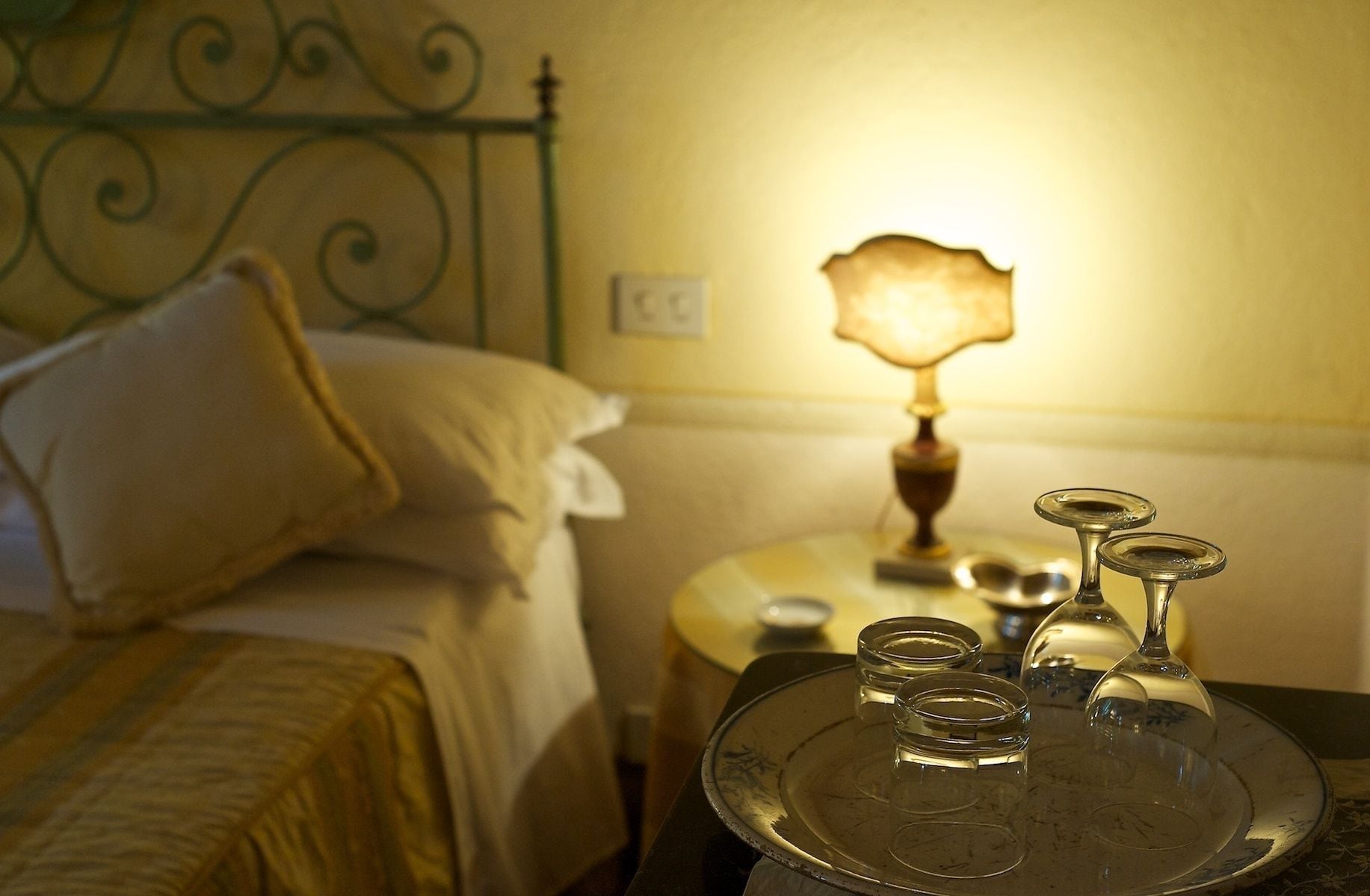 A neatly made bed with a pillow, a bedside table with a lamp, and a tray with glasses and a teapot.