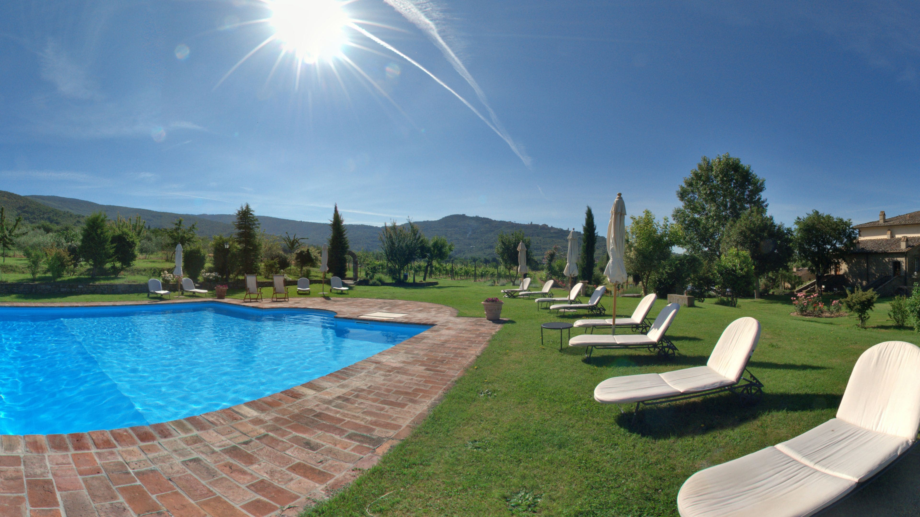 A swimming pool with lounge chairs and umbrellas, surrounded by grass and trees, with a clear sky and mountains in the background.