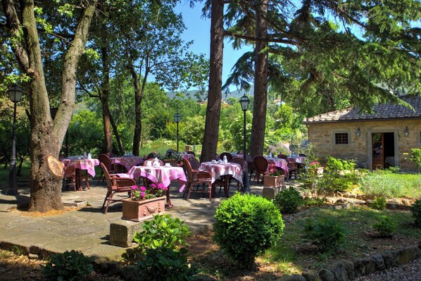 An outdoor dining area with tables and chairs, surrounded by trees and a stone building.
