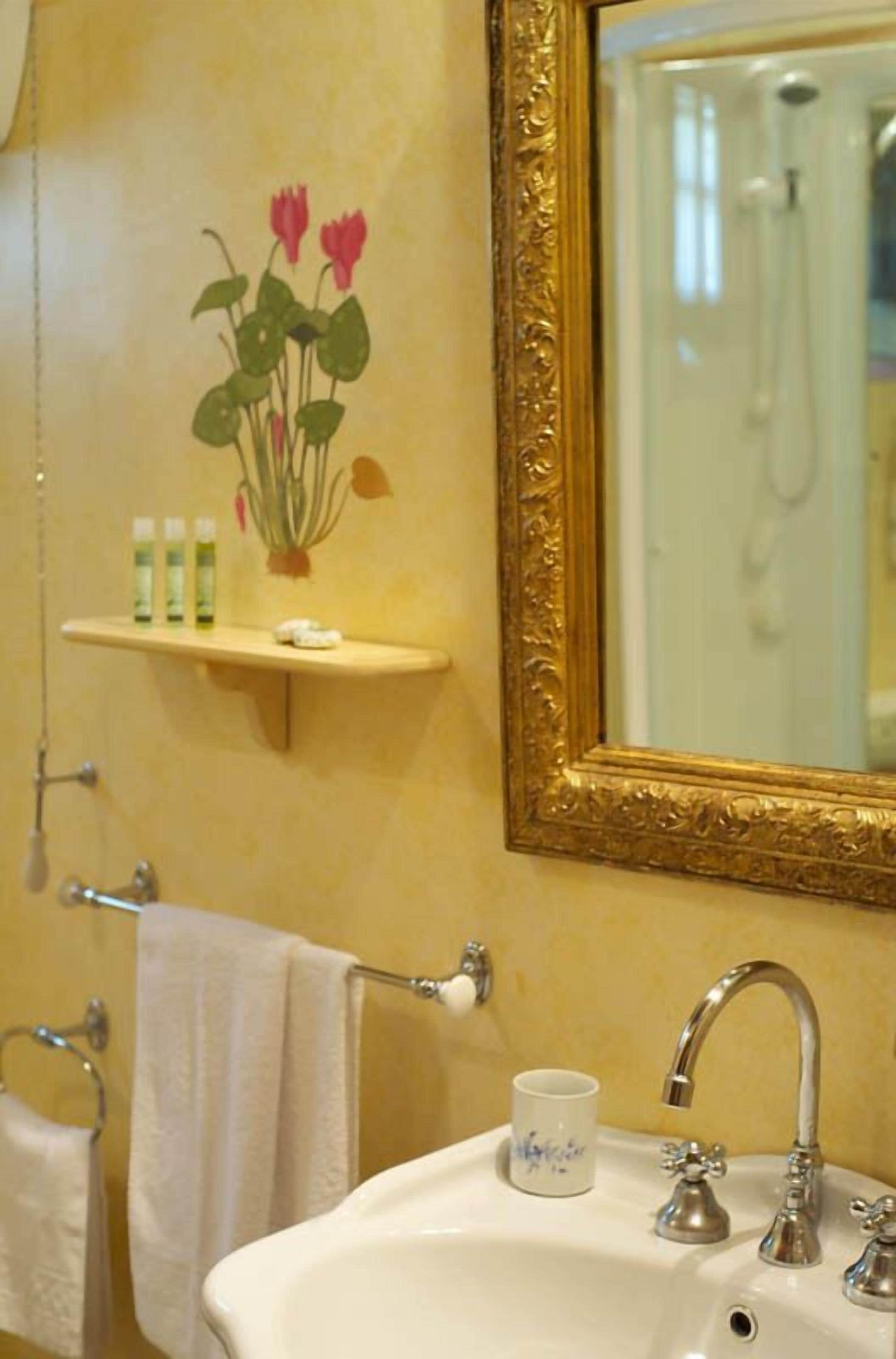 A bathroom with a sink, mirror, and towel rack. There is a shelf with toiletries and a decorative flower painting on the wall.