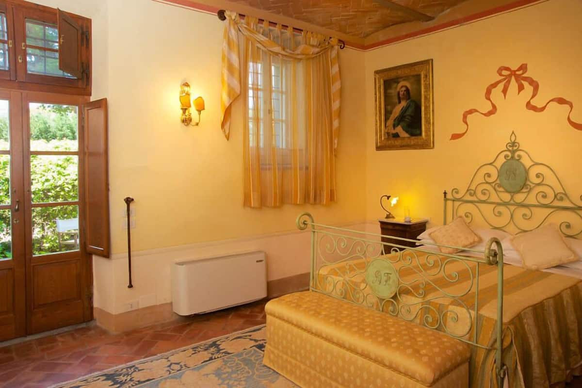 Un dormitorio con cama, mesita de noche, lámpara de pared, radiador y ventana con cortinas.