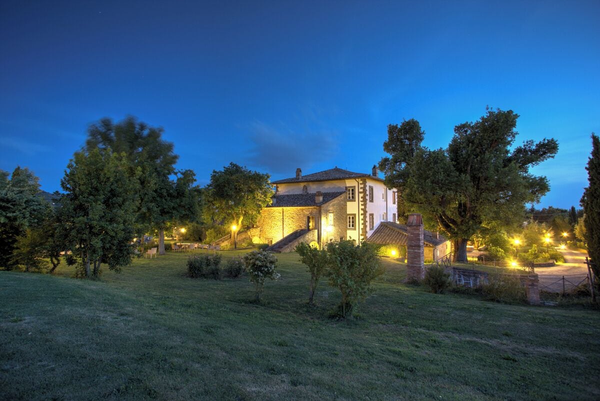 A two-story stone house with a sloped roof, surrounded by a well-maintained lawn and trees.
