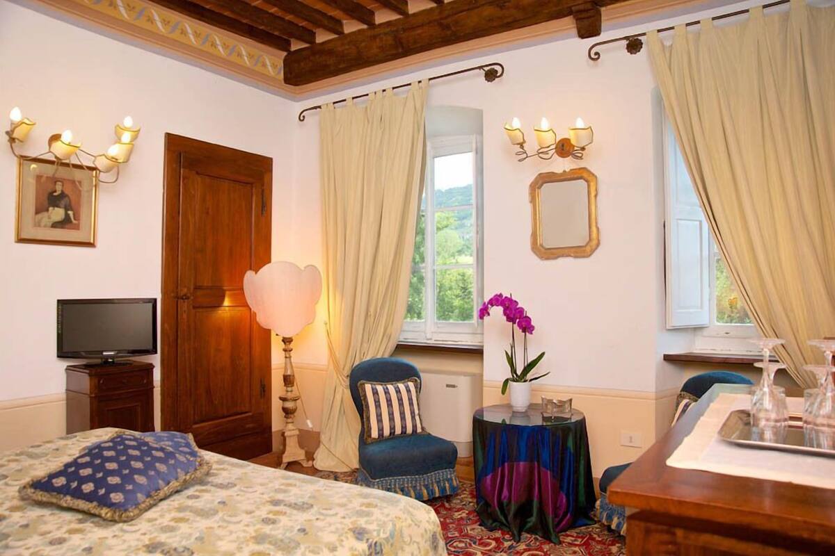 Habitación de hotel con cama, una silla, una mesa con un florero y televisor, además de un ventanal con cortinas.