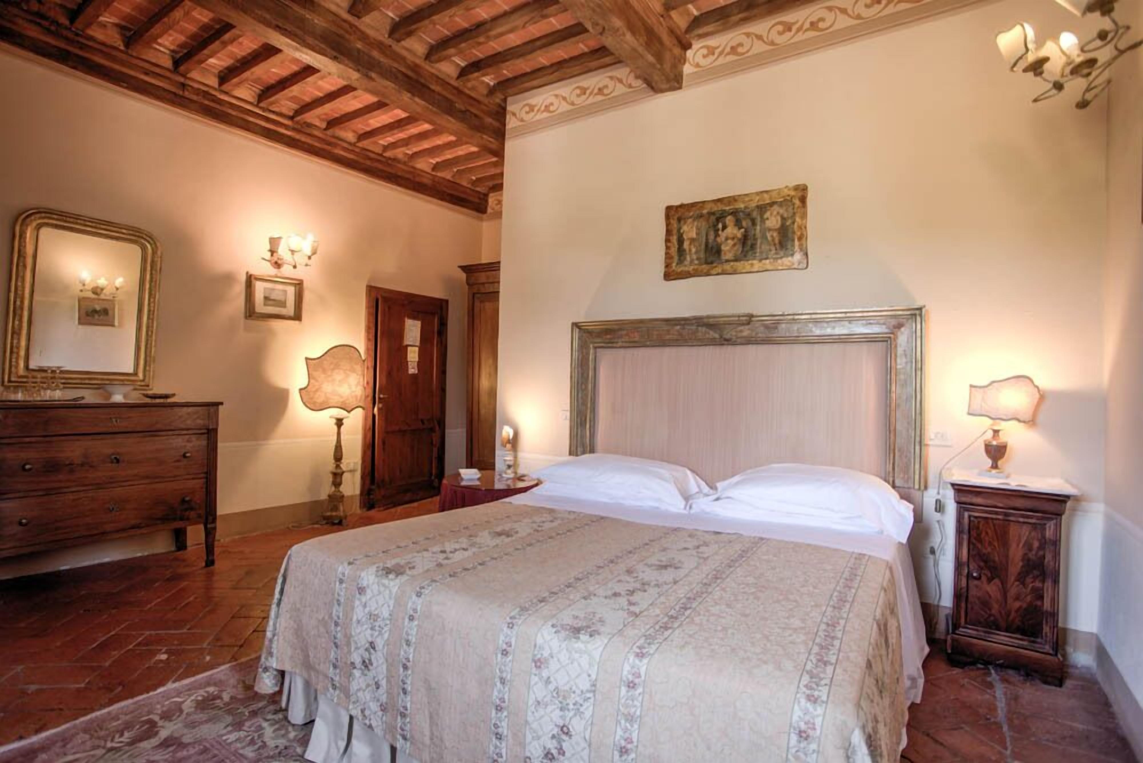 A bedroom with a wooden ceiling, a bed with a floral bedspread, a wooden dresser, a nightstand with a lamp, and a framed picture on the wall.