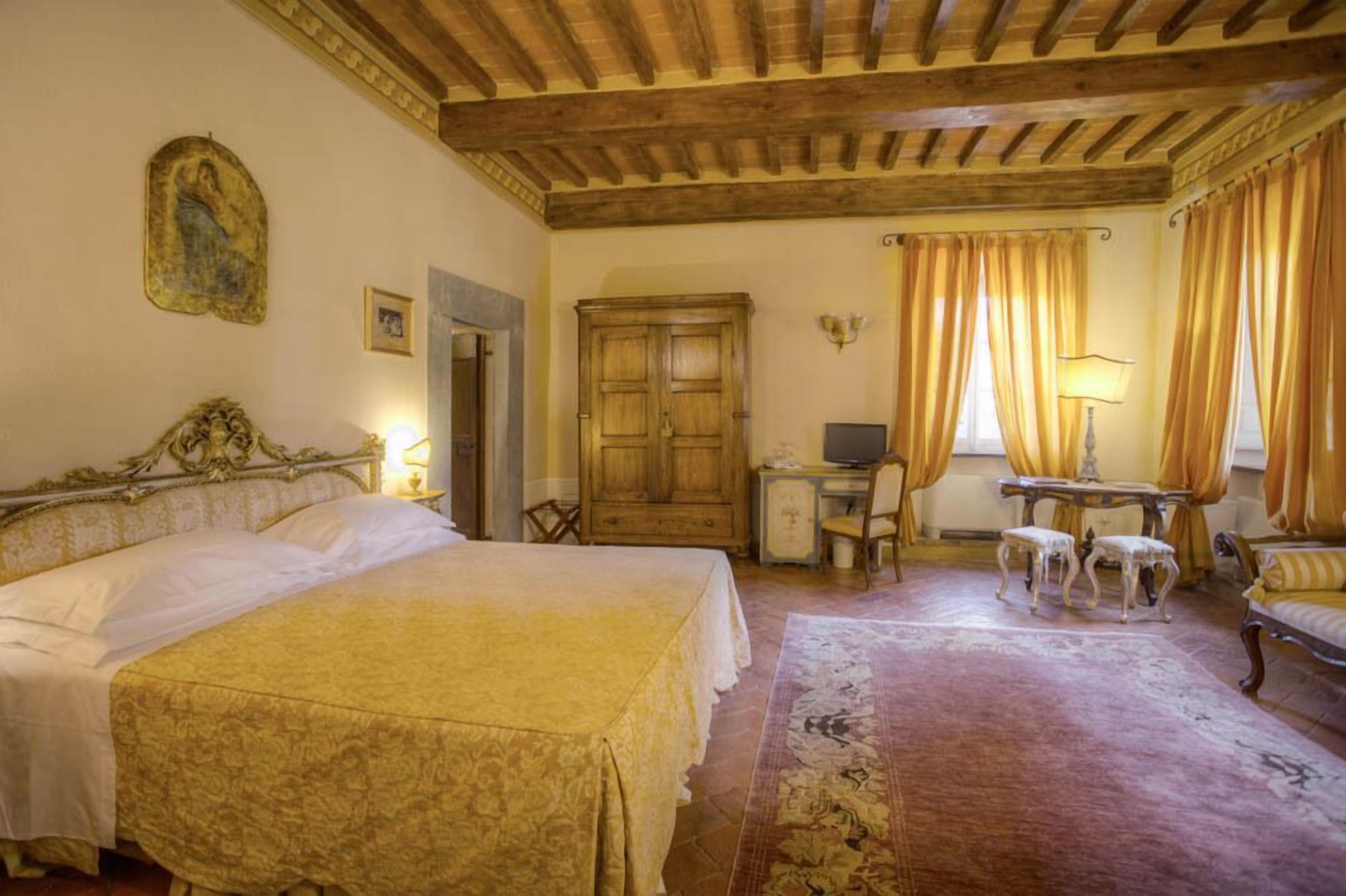 A bedroom with a large bed, a wooden wardrobe, a desk with a chair, and a small table with two stools.