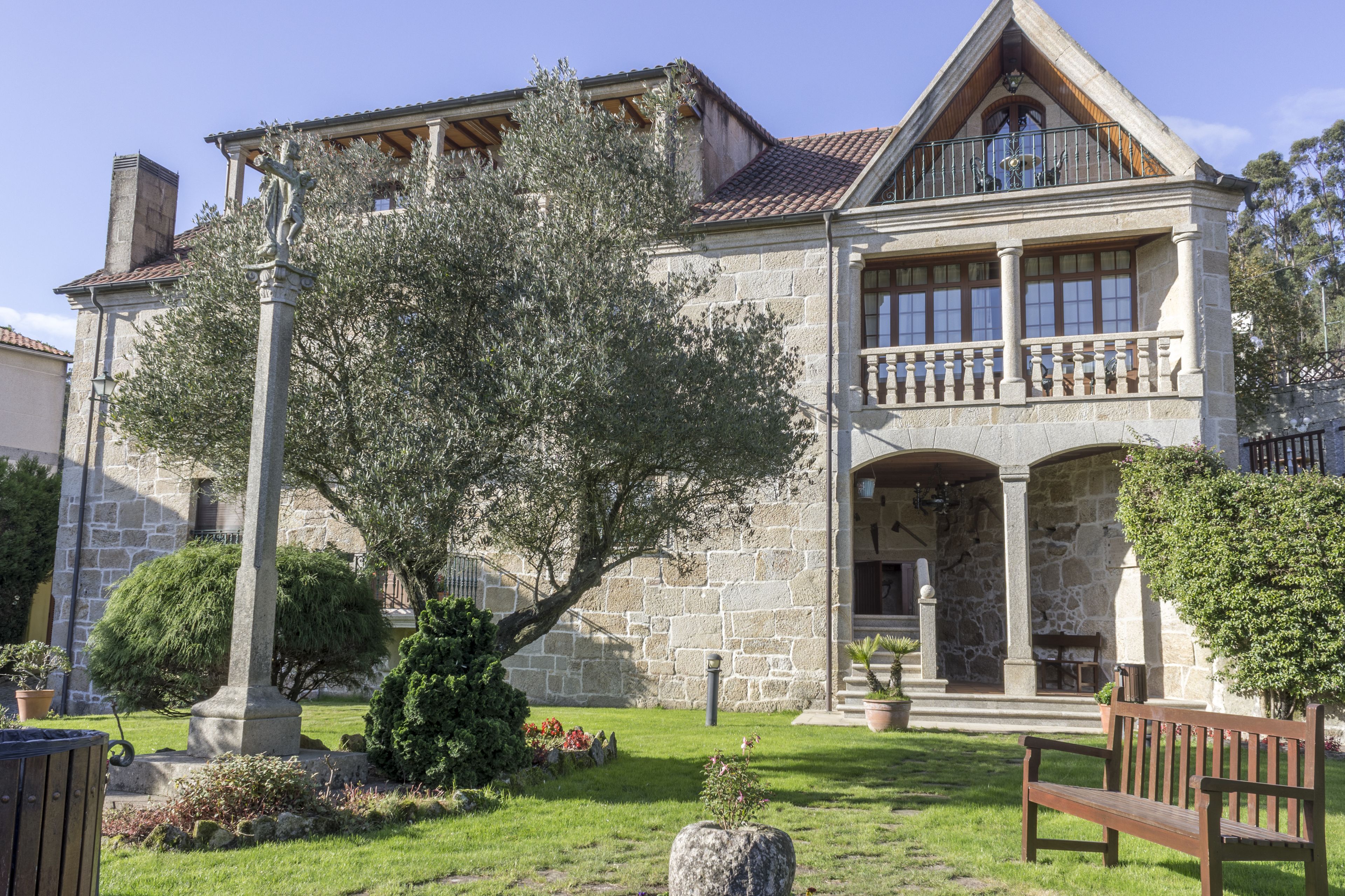 A stone building with a large tree in the front yard, a balcony, and a bench.