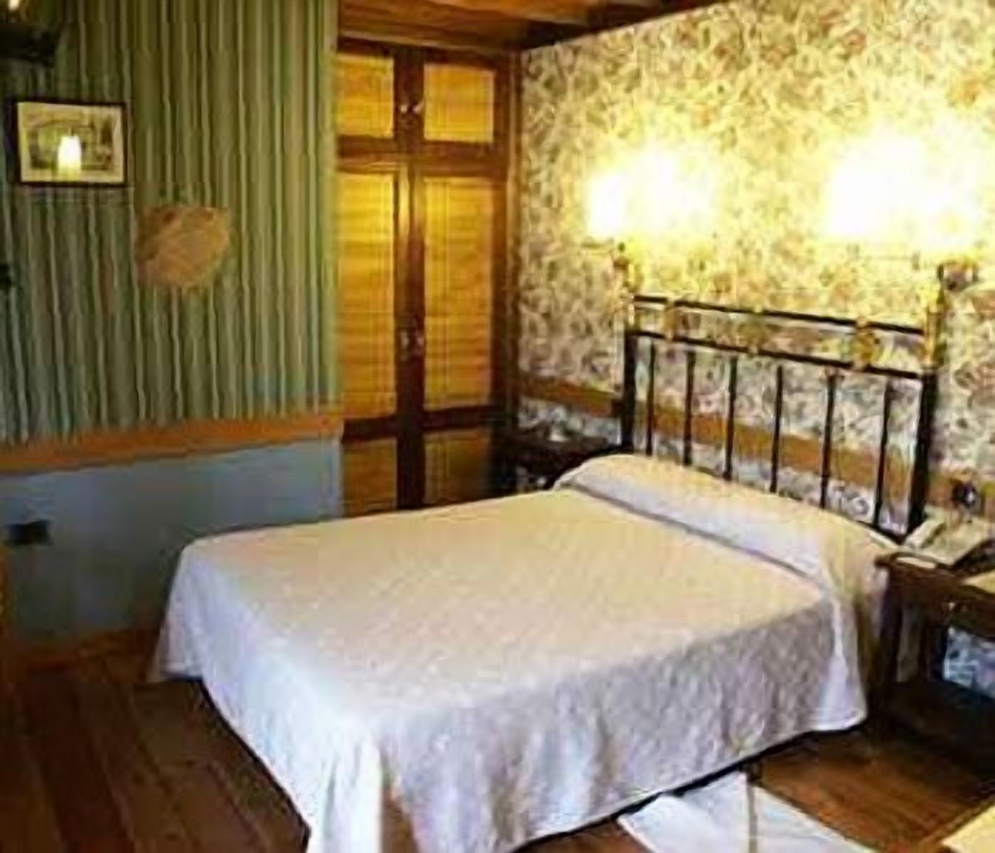 A bedroom with a bed, wooden floor, a window with curtains, and a wall with a patterned wallpaper.