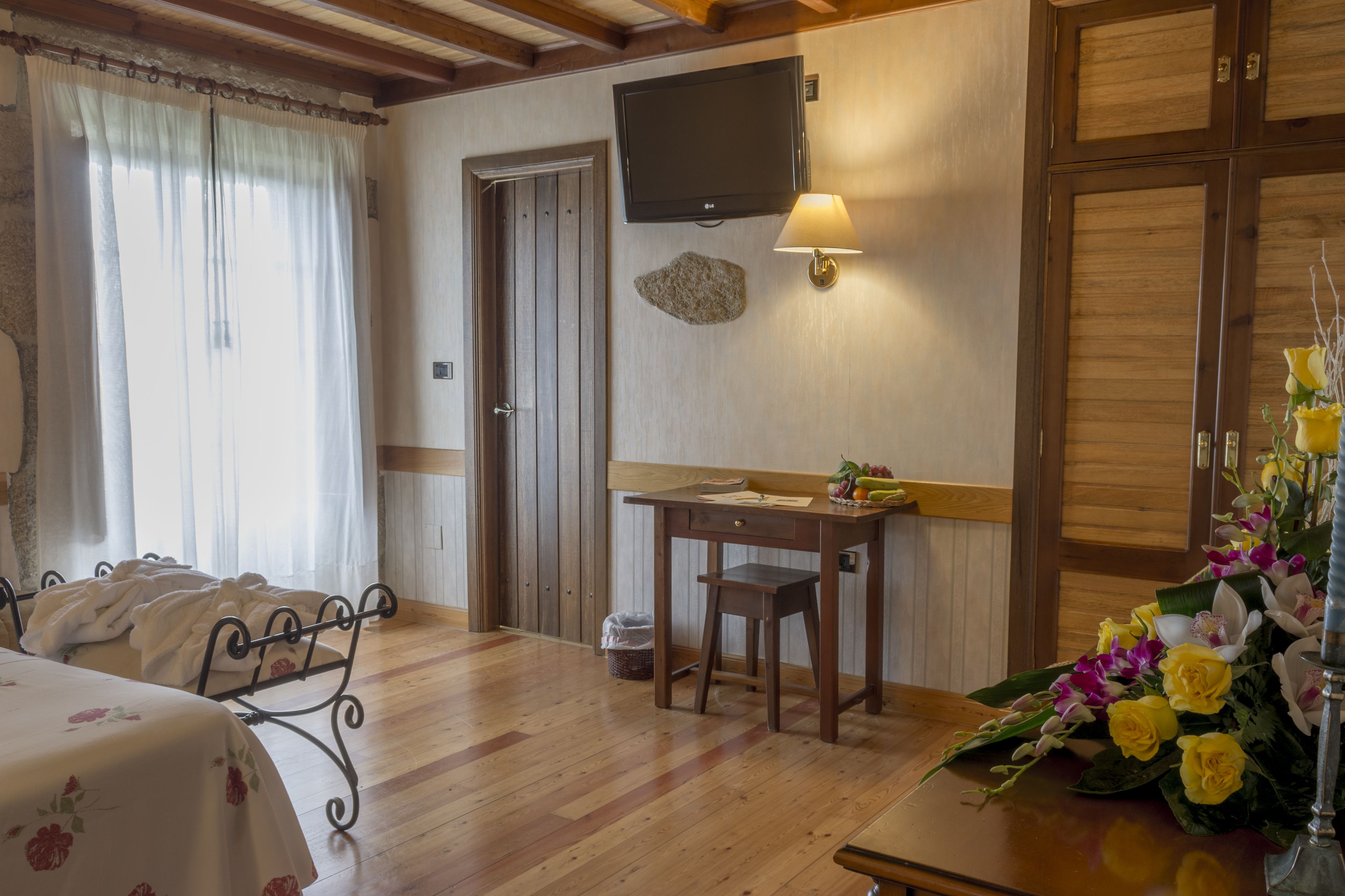 Habitación de hotel con piso de madera, cama con colcha estampada, escritorio con lámpara, televisor montado en la pared y un jarrón de flores sobre el escritorio.