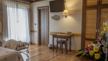 Habitación de hotel con piso de madera, cama con colcha estampada, escritorio con lámpara, televisor montado en la pared y un jarrón de flores sobre el escritorio.
