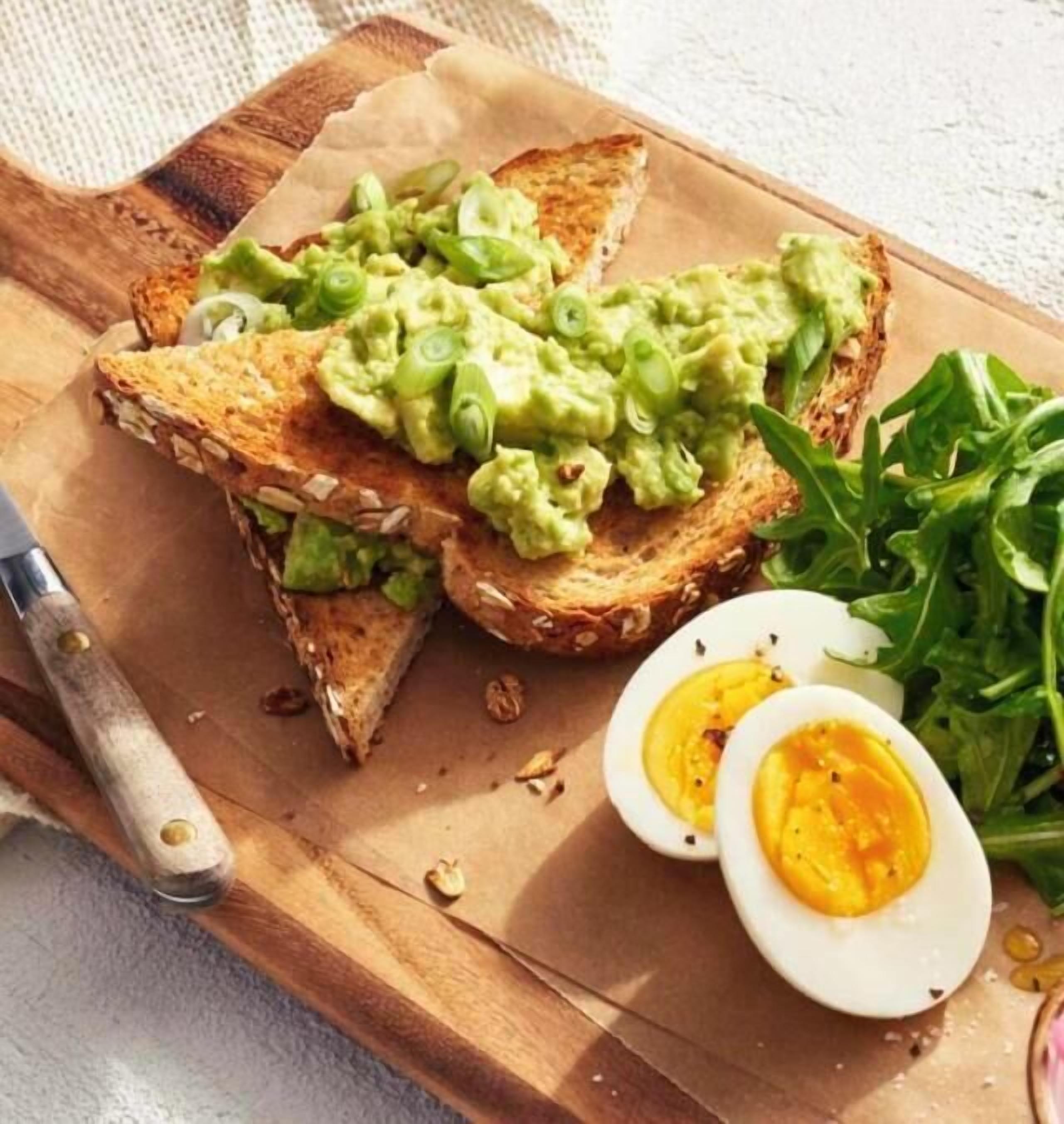 Un sándwich sobre una tabla de madera, un cuchillo, un bol de ensalada y un plato con un huevo cocido.