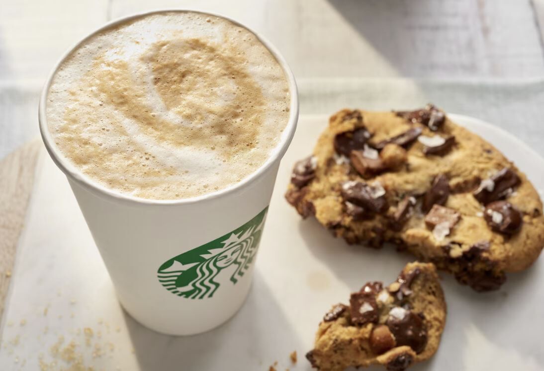 A Starbucks cup with a frothy beverage and a cookie with chocolate chips.